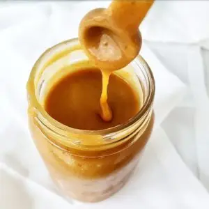 A glass jar of warm, golden-brown Pumpkin Caramel Sauce drizzled on a stack of pancakes.