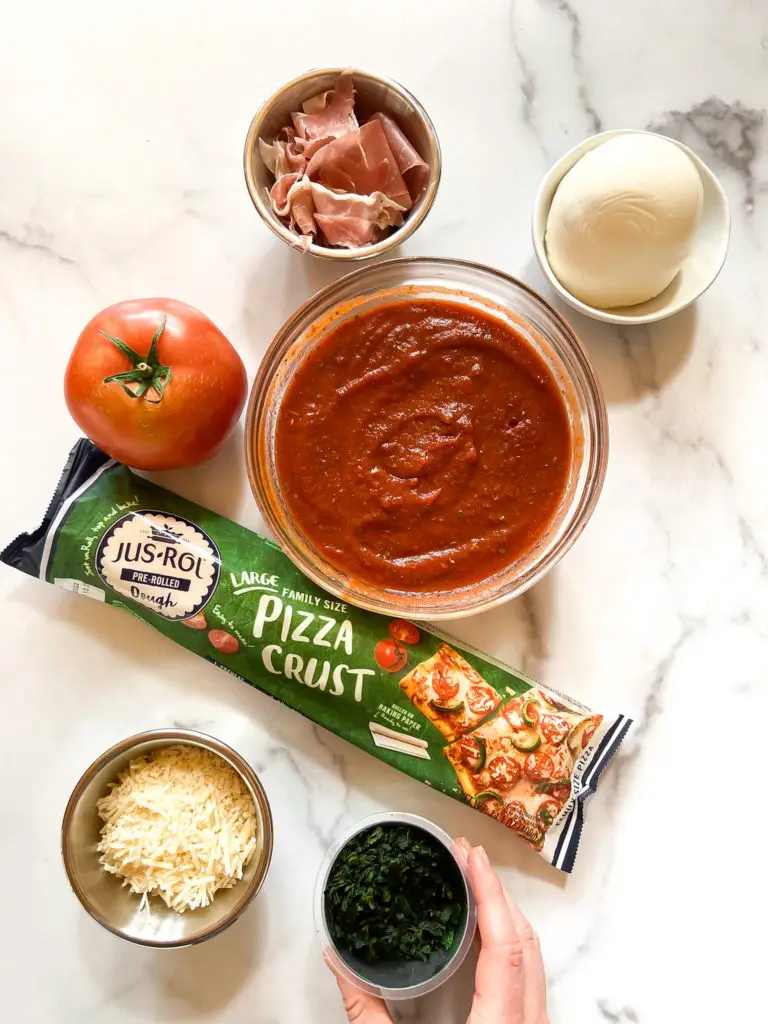 A bowl of vibrant, red homemade pizza sauce with fresh basil leaves on top, sitting next to a wooden spoon.