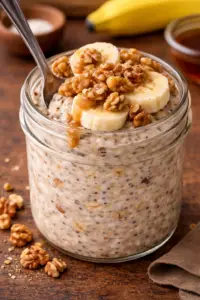Cottage cheese banana bread overnight oats in a mason jar topped with banana slices, walnuts, and maple syrup with a spoon.