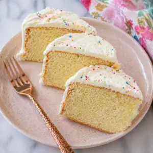 Eggless vanilla cake slices on a pink ceramic plate with rose gold fork, soft moist crumb and light buttercream frosting with sprinkles
