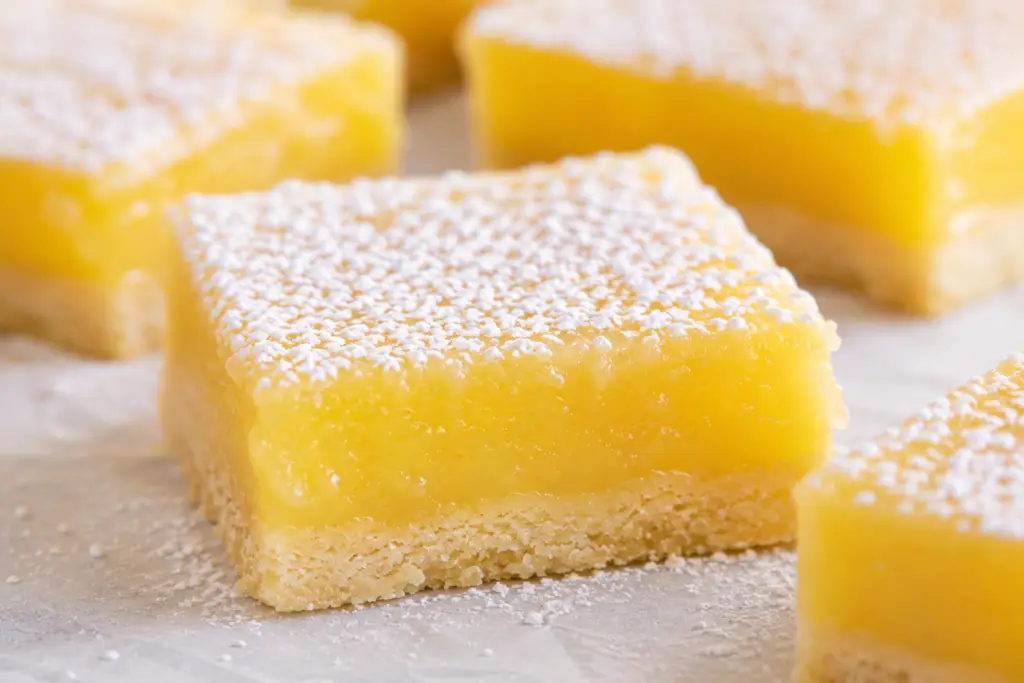 Close-up of bakery-style lemon bars dusted with powdered sugar featuring thick creamy lemon filling and a buttery shortbread crust with fresh lemons in the background.