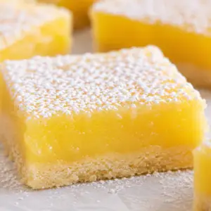 lemon bars recipe- The Best Lemon Bars Recipe (Bakery-Style, Bright, Buttery, and Perfectly Set) Close-up of bakery-style lemon bars dusted with powdered sugar featuring thick creamy lemon filling and a buttery shortbread crust with fresh lemons in the background.