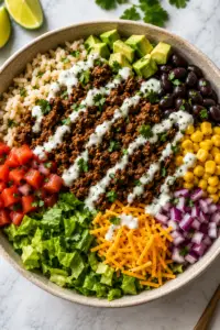 Low-cholesterol ground beef burrito bowl topped with cilantro lime brown rice, seasoned taco beef, black beans, corn, diced tomatoes, red onion, romaine lettuce, avocado, shredded cheddar cheese, and a creamy Greek yogurt drizzle.