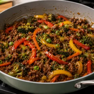Asian ground beef stir fry with broccoli, bell peppers, carrots, and savory stir fry sauce served in a sage green skillet on a stovetop.