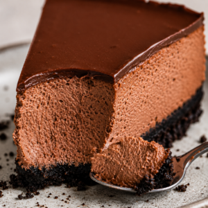 Close-up slice of chocolate cheesecake with a spoonful removed, revealing the creamy chocolate filling and Oreo cookie crust beneath a smooth ganache topping.