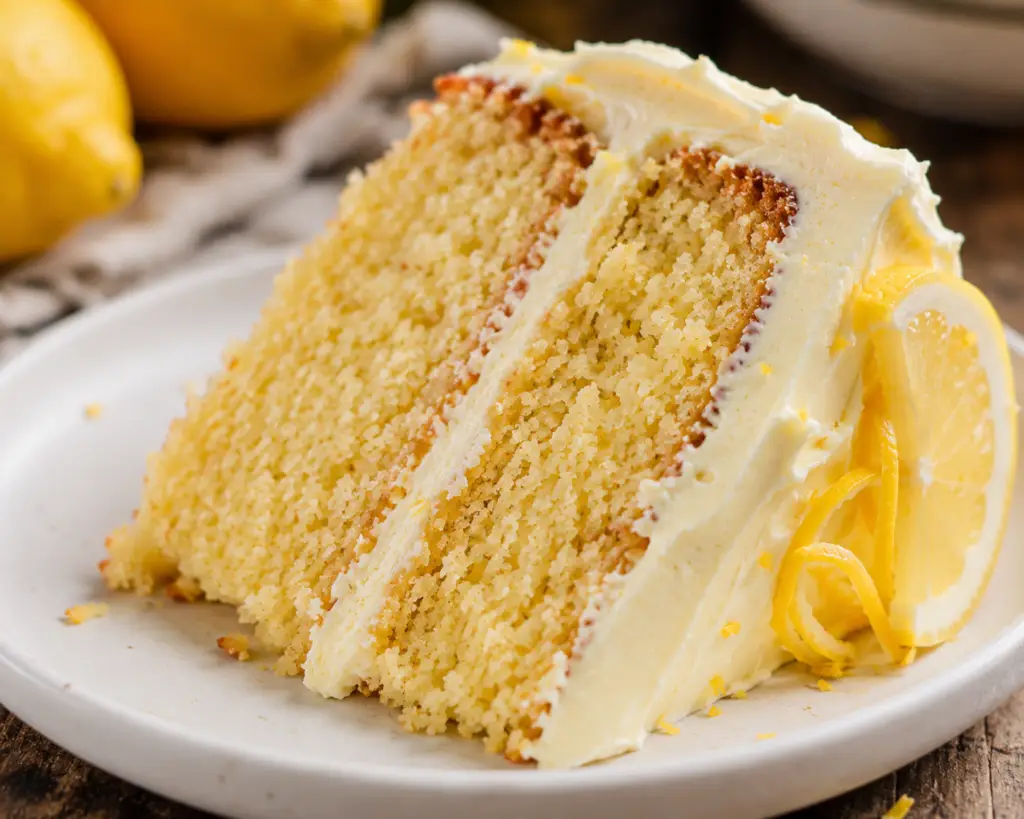 Close-up of a rustic slice of homemade lemon layer cake with lemon cream cheese frosting on a white plate, showing a soft, moist crumb and fresh lemon garnish.