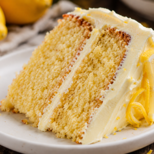 Close-up of a rustic slice of homemade lemon layer cake with lemon cream cheese frosting on a white plate, showing a soft, moist crumb and fresh lemon garnish.