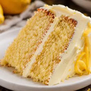 Close-up of a rustic slice of homemade lemon layer cake with lemon cream cheese frosting on a white plate, showing a soft, moist crumb and fresh lemon garnish.