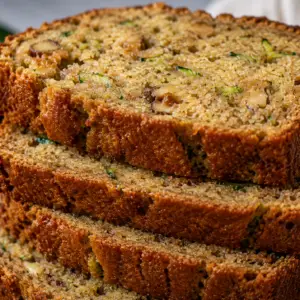 Stack of thick slices of moist homemade zucchini bread with walnuts and shredded zucchini, showing a soft cinnamon-spiced crumb and golden crust.