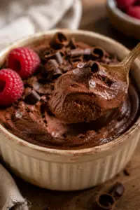 Vintage gold spoon lifting a creamy bite of cottage cheese chocolate mousse from a rustic ceramic ramekin with chocolate curls and fresh raspberries.