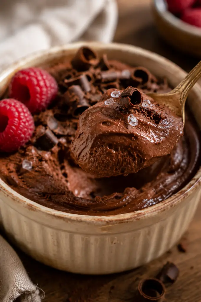 Vintage gold spoon lifting a creamy bite of cottage cheese chocolate mousse from a rustic ceramic ramekin with chocolate curls and fresh raspberries.