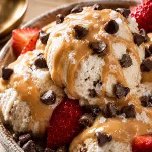 Close-up of creamy homemade cottage cheese ice cream topped with peanut butter drizzle, mini chocolate chips, flaky sea salt, and fresh strawberries in a rustic ceramic bowl.
