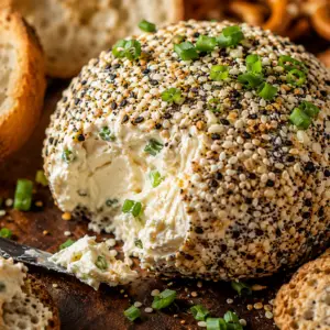 everything bagel cheese ball- Everything Bagel Cheese Ball Close-up of a rustic everything bagel cheese ball coated in everything seasoning with a scoop removed to reveal the creamy interior, surrounded by crackers and pretzels.