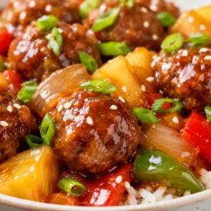 Close-up of homemade sweet and sour meatballs coated in glossy pineapple sauce with bell peppers, onions, pineapple chunks, sesame seeds, and green onions.
