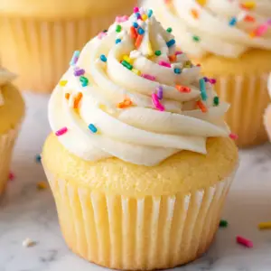 vanilla cupcakes- Vanilla Cupcakes Close-up of a homemade vanilla cupcake topped with a tall swirl of creamy vanilla buttercream frosting and colorful rainbow sprinkles.